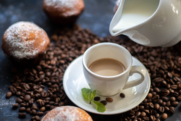 Cup of coffee on a dark background. White milkman. Milk is pouring from it into a cup. On a saucer there are mint leaves, brown grains are scattered nearby, cupcakes cooked at home lie. 