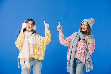 attractive and smiling women in sweaters and scarves pointing with fingers isolated on blue