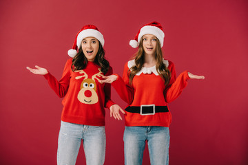 attractive and shocked women in sweaters and santa hats with outstretched hands isolated on red