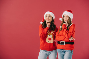 attractive and pensive women in sweaters and santa hats looking away isolated on red