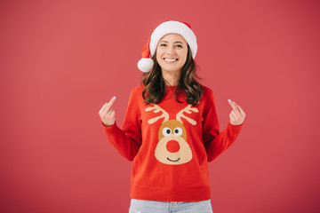 attractive and smiling woman in santa hat and sweater showing middle fingers isolated on red