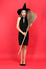 Beautiful woman in halloween costume holding broom on red background