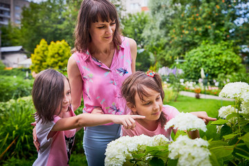 Mother and two funny little girls in green nature park with flowers. Happy sisters and them mom in a summer day