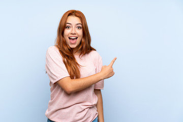 Teenager redhead girl over isolated blue background surprised and pointing side