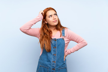 Fototapeta premium Teenager redhead girl with overalls over isolated blue background having doubts and with confuse face expression