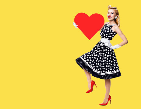 Full body of happy woman holding red heart symbol, dressed in pin up style black dress with white polka dot, isolated over yellow color background