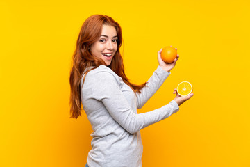 Teenager redhead girl holding an orange over isolated yellow background