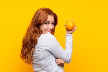 Teenager redhead girl holding an orange over isolated yellow background