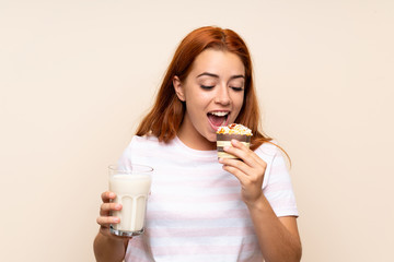 Teenager redhead girl holding a glass of milk and a muffin over isolated background