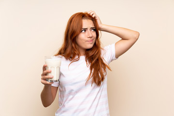 Teenager redhead girl holding a glass of milk over isolated background having doubts and with...