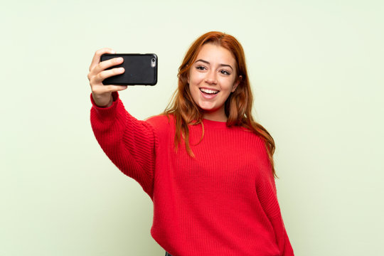 Teenager redhead girl with sweater over isolated green background making selfie with cellphone
