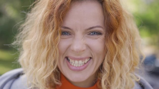Close-up face of cute mature red-haired woman with curly hair smiling happily looking at the camera in the park. Emotions, happiness, good mood