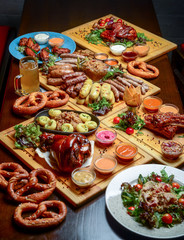 Flat-lay of Oktoberfest dinner table with grilled meat sausages, pretzel pastry, potatoes, cucumber salad, sauces, beers