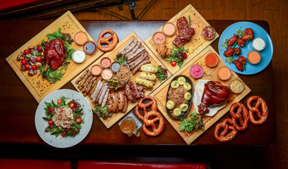 Flat-lay of Oktoberfest dinner table with grilled meat sausages, pretzel pastry, potatoes, cucumber salad, sauces, beers