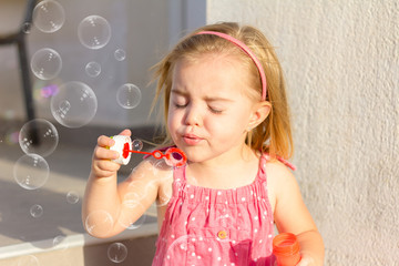 A cute blonde little girl, with a pink dress, blowing soap bubbles