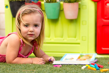A cute blonde little girl laying on the grass and reading a book. There is a colorful toy house on the background and some crayons on the grass.