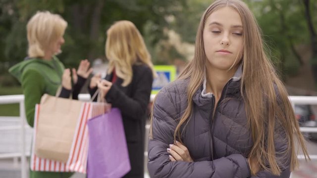 Young sad woman feeling envy the foreground while her two beautiful blond girlfriends standing on the street showing each other shopping bags