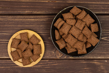 Lot of whole crispy brown cereal pillow in dark ceramic bowl on bamboo coaster flatlay on brown wood