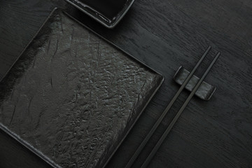 Black chopsticks on chopsticks rest, empty square slate plate and bowl for soy sauce on black wooden table. Flat lay. Mock-up. Asian food concept. Copy space