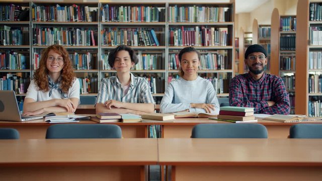 Multi-ethnic group of young people girls and guys students are sitting at desk in library smiling looking at camera. Lifestyle, education and youth concept.