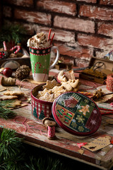 homemade delicious gingerbread cookies in vintage gift round metal box with christmas ornaments on wooden rustic red table with cup of coffee with foam, christmas tree branches opposite brick wall