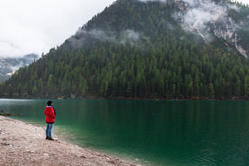 Women with red rain jacket enjoying the peaceful mystic view of a mountain lake with cloudy dramatic mountains in the background on a rainy autumn day.