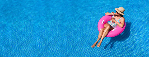 Young woman with sunglasses, hat and swimsuit in a blue pool. Pretty girl on a pink float enjoying the summer while having a cocktail.