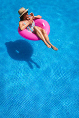Young woman with sunglasses, hat and swimsuit in a blue pool. Pretty girl on a pink float enjoying the summer while having a cocktail.