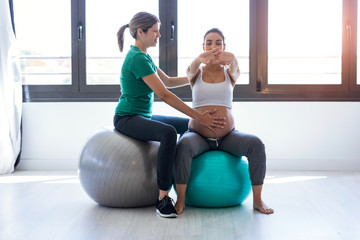 Physiotherapist helping to beautiful pregnant woman for doing pilates exercises with ball preparing...
