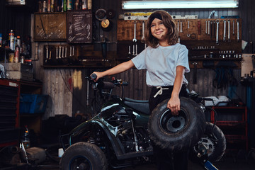 Fototapeta na wymiar Cheerful small girl is posing for photographer with big wheel from broken ATV at auto service.
