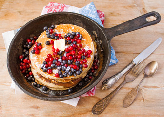 Stack of homemade shrovetide carnival clean eating oat pancakes with powered sugar, blueberries and cowberries in cast iron pan on rustic country table with old silver dishware. Soft focus. Copy space