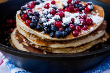 Stack of homemade shrovetide carnival clean eating oat pancakes with powered sugar, blueberries and cowberries in cast iron pan on rustic country table. Soft focus. Copy space