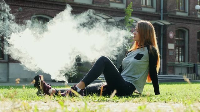 Modern female student smokes vape electronic cigarette and blows steam from mouth while sitting on lawn in campus, rest in summer sunny park
