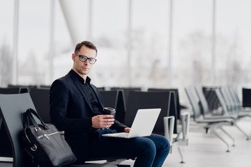Handsome young businessman working on a laptop and drinking a coffee during an expectation of a flight