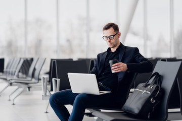 Przystojny młody biznesmen pracuje na laptopie i pije kawę podczas oczekiwania lot - 291778940