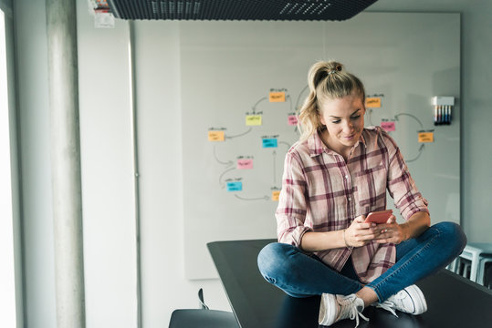 Casual businesswoman sitting on table in office using cell phone