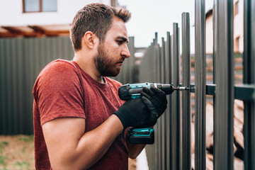 Construction worker using electrical screwdriver and mounting metal elements on fence