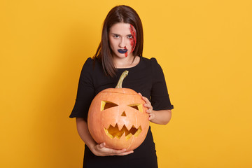 Studio shot of serious girl with angry facial expression standing with pumpkin in hands in studio isolated over yellow background, attractive female with bloody wound on her face, halloween concept.