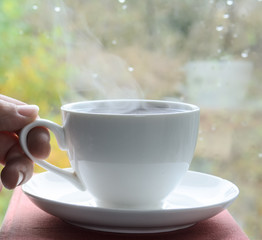 female hand holds a white cup with coffee. A cup is reflected in the window. Concept - autumn warming cup of coffee