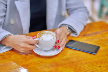 close-up woman hands raise cup with cappuccino. nearby lies the phone on the table