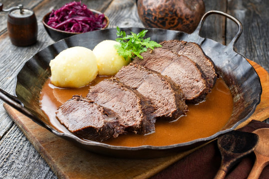 Traditioneller geschmoreter marinierter deutscher Sauerbraten vom Rind mit Kartoffel Kn&ouml;del und Blaukraut in einer w&uuml;rzigen braunen Brauten Sauce als closeup in einer rustikalen Pfanne