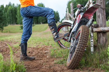 Man rider standing near dirt motorcycle in countryside, steps with boot on bike peg