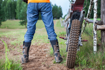 Male rider well shod in protective boots standing near off-road motorcycle on dirt road, rear view