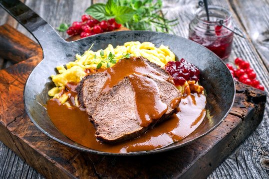 Traditioneller geschmorter marinierter deutscher Sauerbraten vom Rind mit Sp&auml;tzle und Preiselbeeren Marmelade in w&uuml;rziger brauner Bratensauce als closeup in einer rustikalen schmiedeeisernen Pfanne