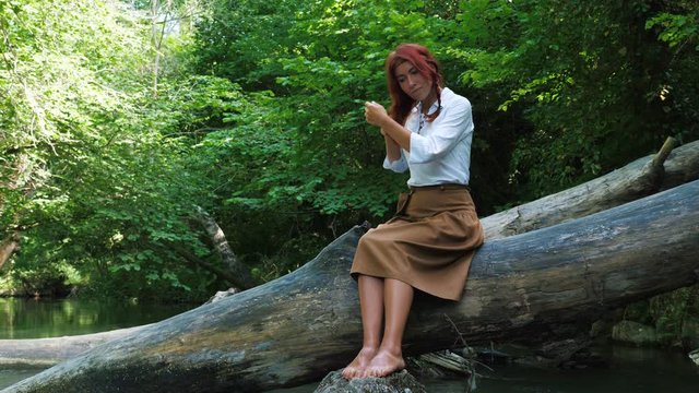 A red-haired girl dressed in a rustic style combs her hair against the background of a forest and a river.