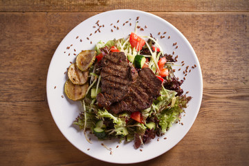 Beef salad: grilled beef steaks with tomatoes, cucumbers, zucchini, lettuce and cabbage, sprinkled with flax seeds. Overhead, horizontal image