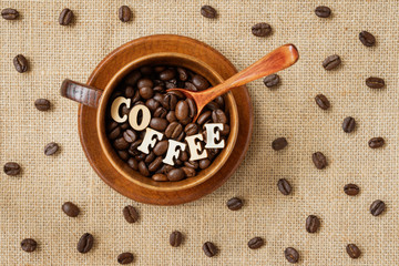 Roasted coffee beans with word coffee in wooden cup and wooden spoon on natural canvas, sackcloth with coffee beans, top view, selective fokus