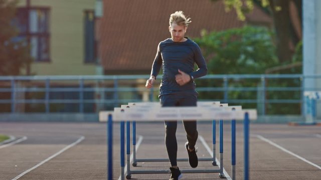 Athletic Fit Man in Grey Shirt and Shorts Hurdling in the Stadium. He is Jumping Over Barriers on a Warm Summer Afternoon. Athlete Doing His Routine Sports Practice. Slow Motion Tracking Shot.