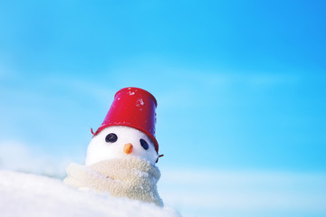 Little snowman made of snow in scarf and red bucket on his head on snow on blue sky background, soft focus. Christmas background