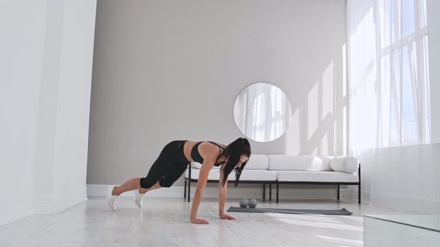 Beautiful slim woman in thirties with fair complexion does morning strenth and aerobic burpee exercises in modern airy room.